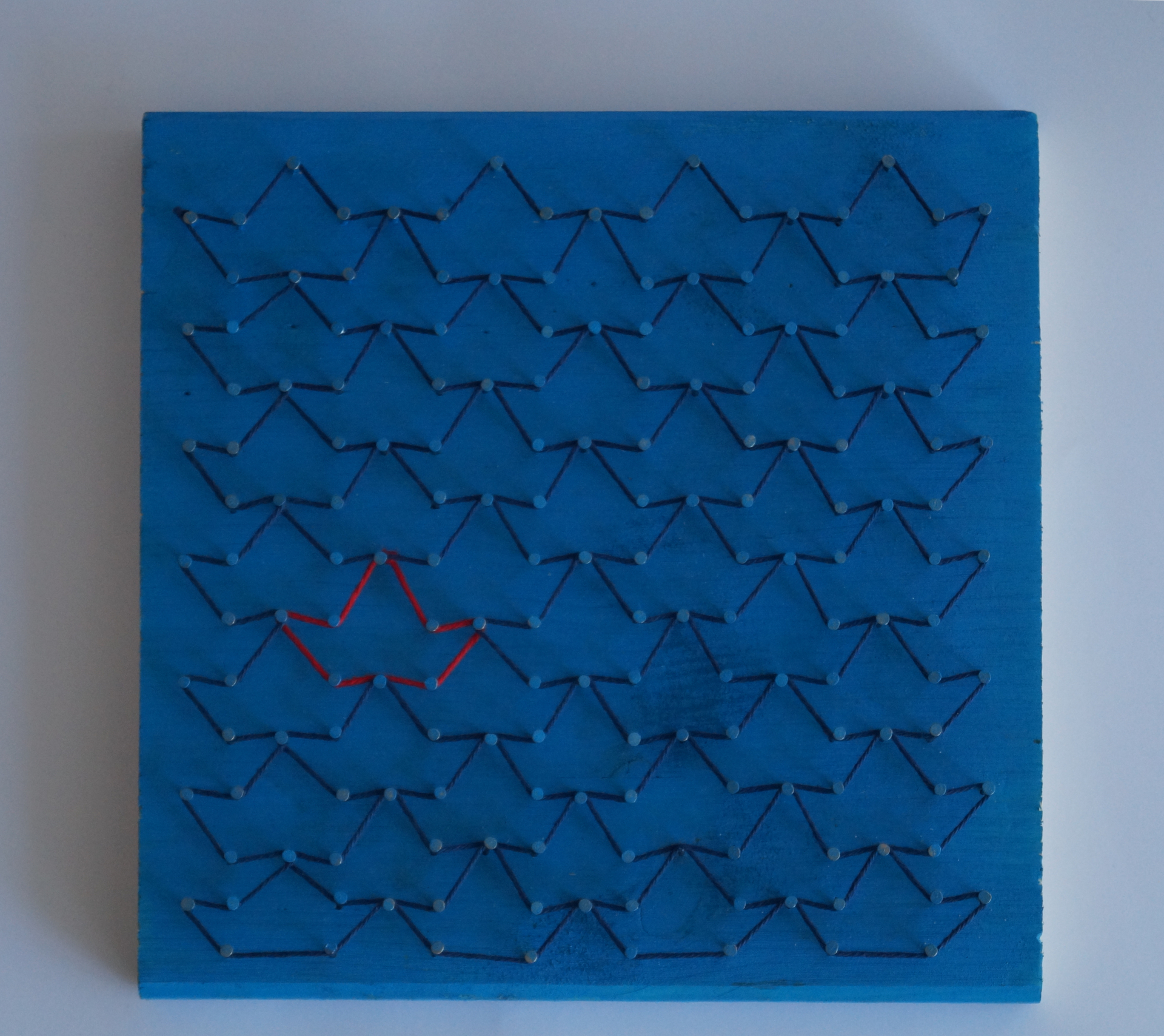 A wooden board painted blue with nails hammered into it that are connected via a dark blue thread. The way the nails are connected reminds of paper boats. One of these is highlighted red.
