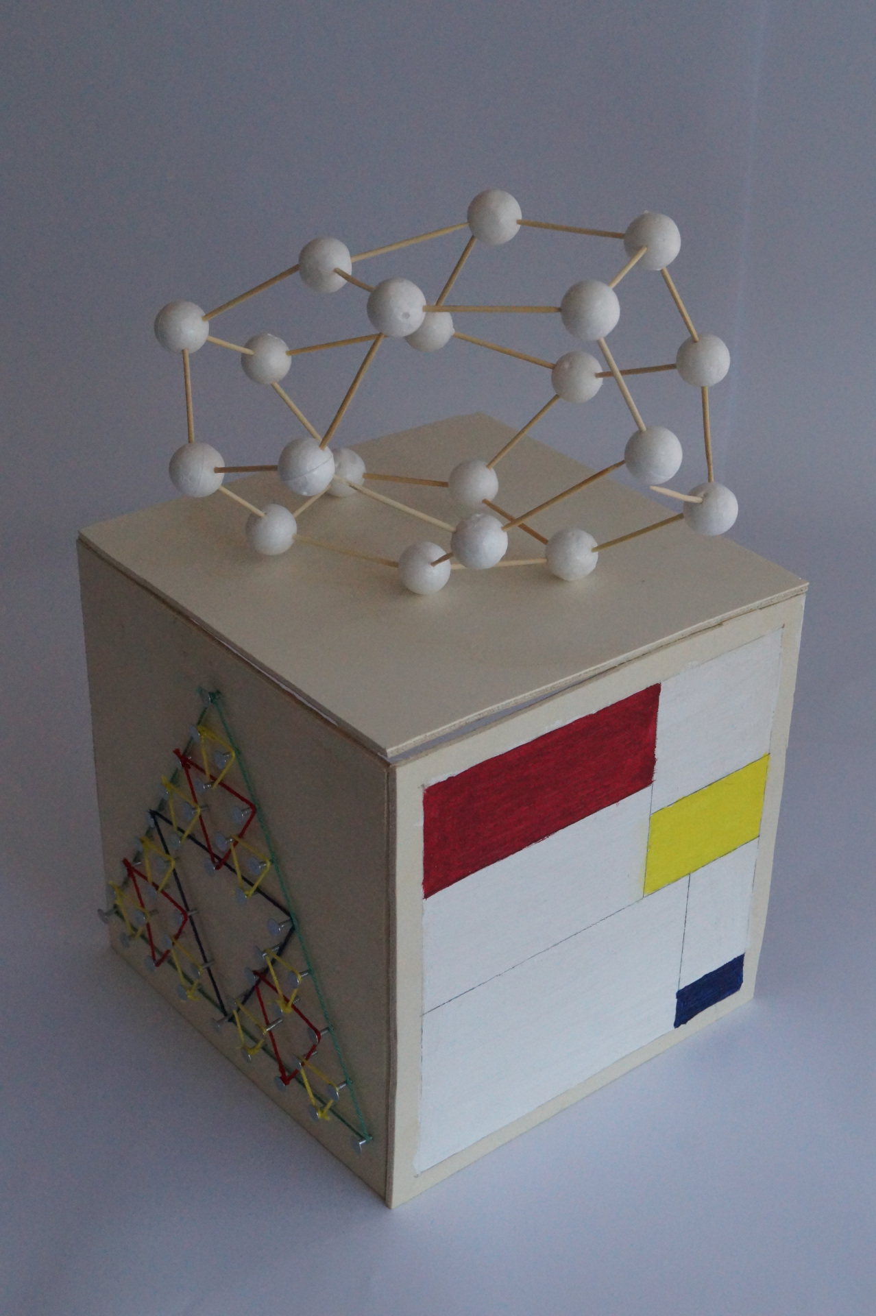 A cube made from plywood. On the left side, a string art Sierpinski triangle is shown. On the right side, a stylized image reminiscent of the work of Piet Mondrian is painted. On top of the cube rests a graph structure made of styrofoam balls, connected via toothpicks.